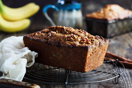 Front side view of a loaf of fresh homemade banana bread cooling on a baker's rack with ingredients nearby. Selective focus on loaf with blurred backgroundの写真素材