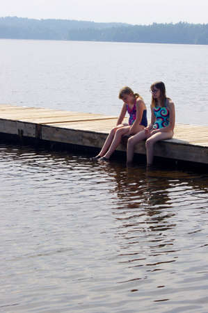 Two young girls sitting in swimsuits on wooden dock in waterの写真素材