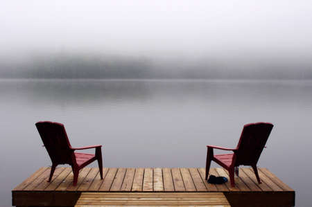two Adirondack chairs on wooden deck at edge of lake in fog with copy spaceの写真素材