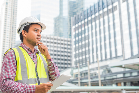 Young handsome asian civil engineer holding tablet looking forward and thinking, planning on construction building background. Evolution construction concept.の写真素材