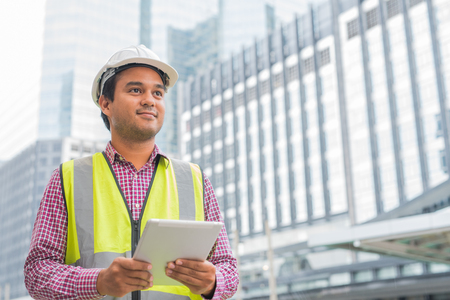 Young handsome asian civil engineer holding tablet looking forward and thinking, planning on construction building background.の写真素材