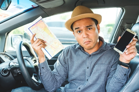 Bad navigation. Young man having a problem with orientation on the road during his journey with a car.He holding a map and smartphone.Get lost Traveling and transportation conceptの写真素材