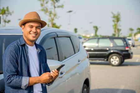 An Asian man is standing using a smartphone beside a car.