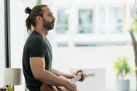 Handsome beard man playing guitar. Hipster man wearing green shirt holding guitar sitting in house.