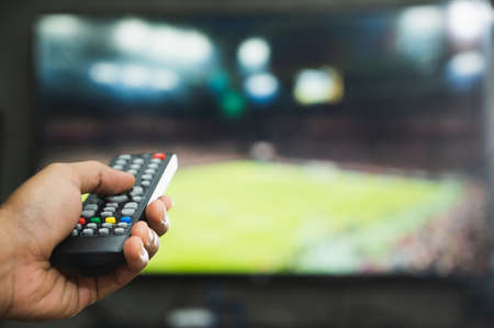 Young man holding television remote control watching football program. Hands pointing to tv screen set and turning it on or off select channel watching tv in the living room relax.の写真素材