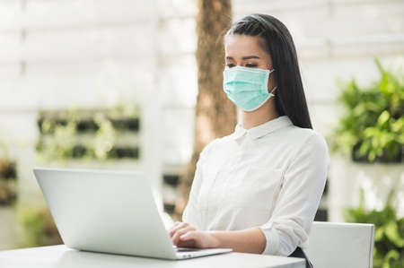 Beautiful asian woman wearing protection mask work from home in the morning. Young asian female with laptop stay at home.の写真素材