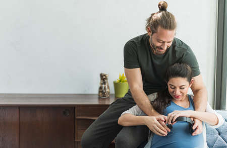 Beard man and his pregnant wife are hugging and giving headphone play the music for they baby.