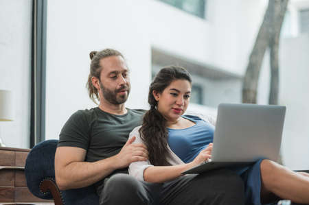 Beard man and his pregnant wife watching movie on laptop feeling happy while sitting on sofa in the...