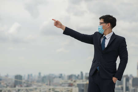 Young handsome asian businessman standing wearing mask of protect the pollution and virus and pointing the finger on building urban cities background.の写真素材