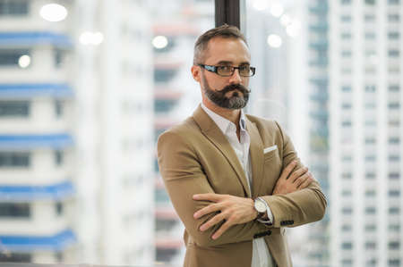 Young handsome business bearded man standing near window with arms crossed in office work place.の写真素材