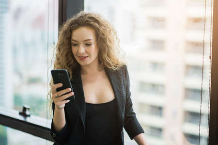 Beautiful sexy woman using smartphone standing near window in office.