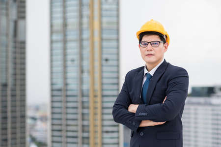 Young handsome asian civil engineer standing wearing helmet yellow hard hat in business suite on building urban cities background.の写真素材