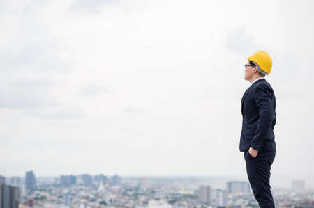 Young handsome asian civil engineer standing wearing helmet yellow hard hat in business suite on building urban cities background.の写真素材