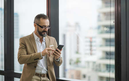 Young handsome business bearded man standing post near window using smartphone in office work place.
