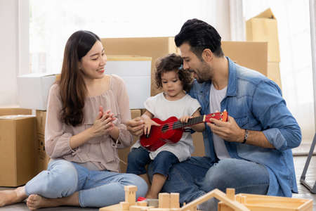 Parents and daughters play with ukulele sitting on the floor in the living room at home. The family just moved to a new house. Happy moment Multi-ethnic dad mom and child.の写真素材