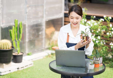 Beautiful Asian young woman greenhouse worker selling Small tree on social media live stream with laptop. Landscaping small plants and decorating the garden in the house. Gardener working house plant.の写真素材
