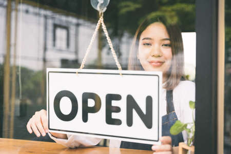 Young asian girl Flip the open label to welcome customer and open the coffee shop in morning. Female asian Barista open the coffee cafe at doorway with reflection in mirror.の写真素材