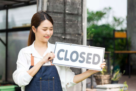 The owner of the tree shop holds a sign close. Her shop has closed. Cute asian gardener small business close her shop selling small tree for decoration in greenhouse.の写真素材