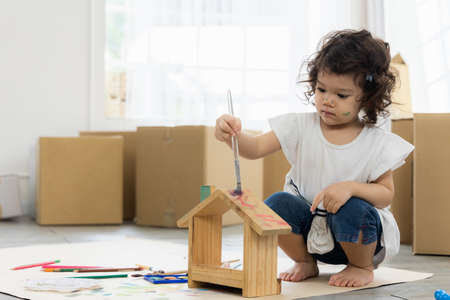 Cute daughter painting coloring with watercolor on the wooden house mock up. Sitting on the floor in living room. Happy kid moment in the house. Art Activity in kindergarten.の写真素材