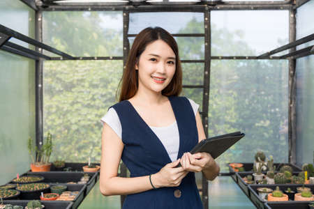 Young cute asian woman working in house plant. Asian female gardener using a tablet to record and capture the growth of trees in greenhouse. Startup small business work from home concept.の写真素材
