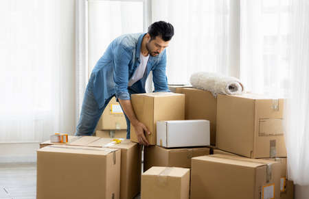 Young man just moving new house. He unpacking parcel box and arranging on the floor in the living...