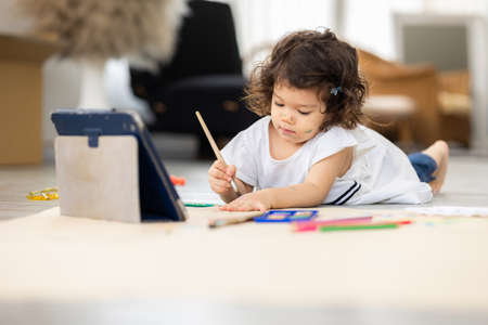 Cute daughter painting coloring with watercolor on the large paper Learning by tablet. Sitting on the floor in living room. Happy kid moment in the house. Art Activity in kindergarten.の写真素材