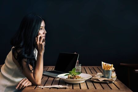 Asian woman using a smart phone for a call at the restaurant. Laptop and healthy dish on the table. Black wall with blankの写真素材