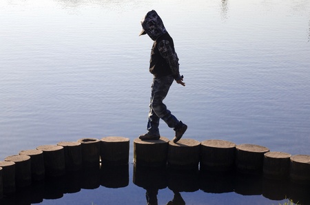 The boy in a jacket with a hood goes, having hung a head, on a construction from logs on river bankの写真素材