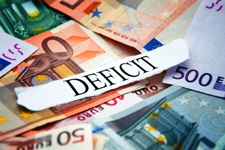 Word "Deficit" printed on a piece of paper and placed above the pile of euro banknotesの写真素材