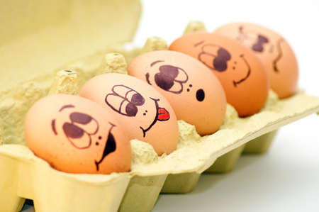 Group of fresh eggs with drawn faces depicting various emotions arranged in a cardboard egg carton against white.の写真素材