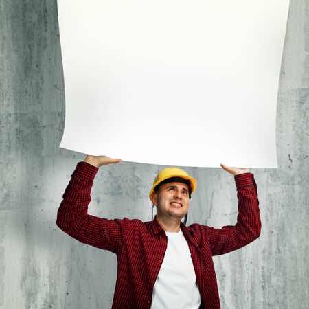Construction worker with yellow hard hat in red shirt holding a whiteboard above his head.の写真素材