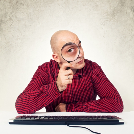 Bald yopung adult man sitting at the table with keyboard holding magnifying glass in front of the computer, internet search conceptの写真素材