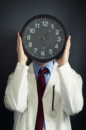 Doctor is working overtime, holding a black clock in front pf his head. Overtime work, not enough time, time pressure, deadline concept.の写真素材