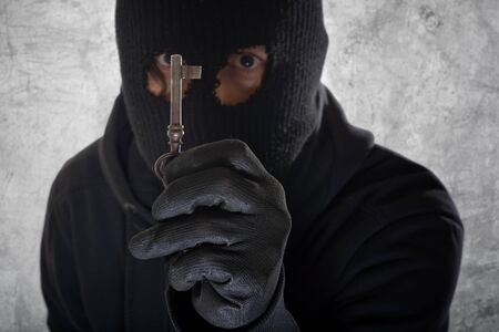 Burglar concept, thief with balaclava caught in front of the grunge concrete wall with an old vintage rusty house key.の写真素材