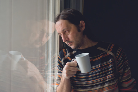 Sad alone mid adult man standing by the window and drinking coffee, looking somewhere in the distance.の写真素材