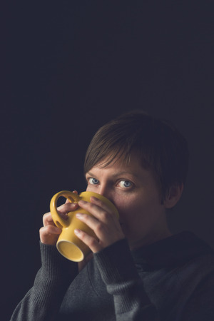 Beautiful caucasian adult woman drinking cup of coffee in dark room. Selective focus with shallow depth of field.の写真素材