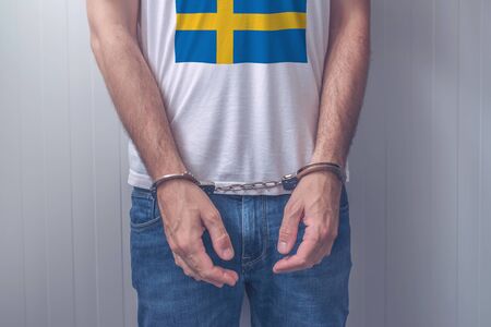 Arrested man with cuffed hands wearing shirt with Swedish flag. Unrecognizable male person in jeans with handcuffs held in police station for being suspected of a crime.の写真素材
