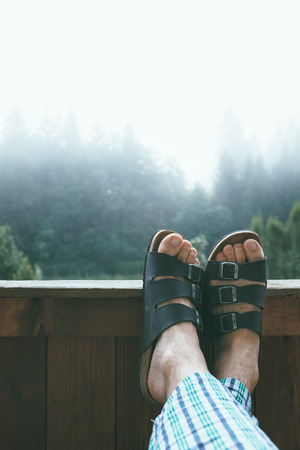 Man enjoying misty morning at mountain hotel balcony, crossed feet in leather slippersの写真素材
