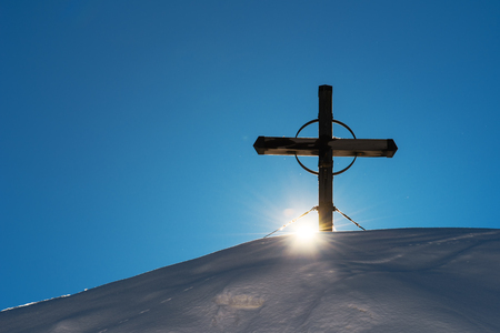 Wooden cross on snow capped mountain hill, setting sun beaming throughの写真素材