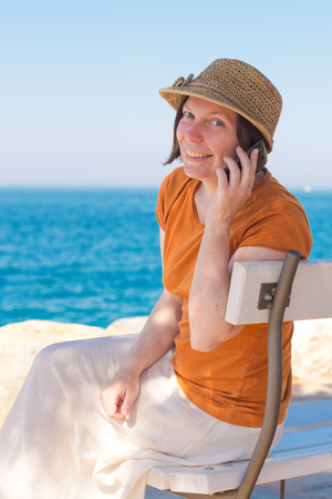 Beautiful adult caucasian woman wearing stylish straw hat is using mobile phone on seaside summer holiday vacationの写真素材