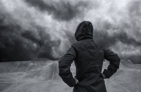 Youth lifestyle concept, unrecognizable hooded person standing in empty futuristic skateboarding park, dramatic sky in background for uncertain and unpredictable futureの写真素材