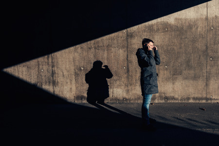 Serious authentic woman with short hair talking on mobile phone on the street in the under passage, winter setting sun casting contrasty shadows on the wallの写真素材