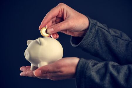 Depositing one euro coin into piggy coin bank for saving, close up of female hand with european...