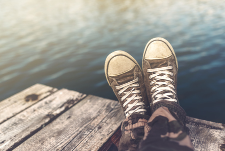 Man with crossed legs relaxing on wooden riverbank pier, casual young guy wearing sneakers sitting by the river in summer afternoon, selective focusの写真素材