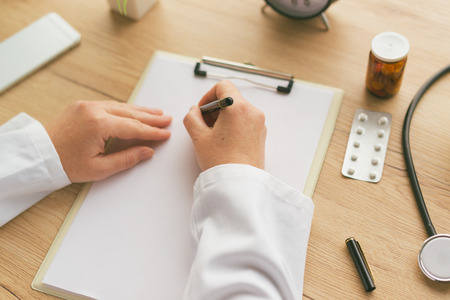 Female doctor writing notes, patient's medical history or medicine prescription on clipboard paper during medical exam in hospital officeの写真素材