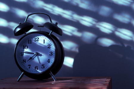Alarm clock on night table in bedroom ticking time in early morning with sunlight and shadows on the wall in backgroundの写真素材