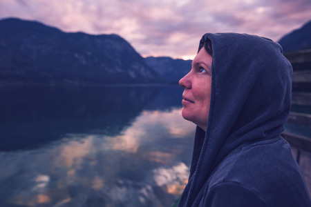 Lonely woman looking at dramatic sky. Female person in hoodie standing alone by the lake, ultra violet toned shadowsの写真素材