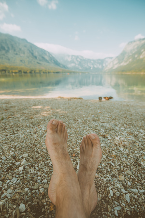 Summer holiday by the lake, barefoot adult caucasian male relaxing on beach pebbles and looking into water and mountains in the distance. Enjoying vacation and nature beauty.の写真素材