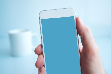 Using smartphone during coffee break in the morning, male hand holding mobile phone with blank screen as copy space in the officeの写真素材
