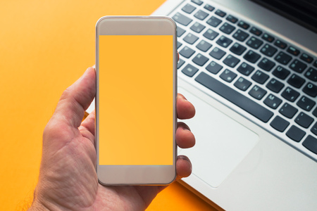 Smart phone in hand with mock up screen, modern laptop computer in background